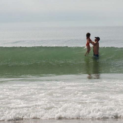 Image of a wave breaking in the ocean with two figures to the right, a father and a daughter. The father, in a baseball cap, is lifting the daughter over the wave as it breaks. The foam of earlier waves are in the foreground. It is a relatively overcast day.