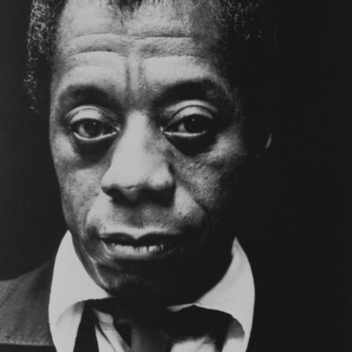 James Baldwin in a black suit with tie and a white shirt looks directly at us.