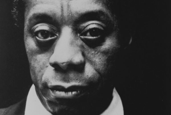 James Baldwin in a black suit with tie and a white shirt looks directly at us.