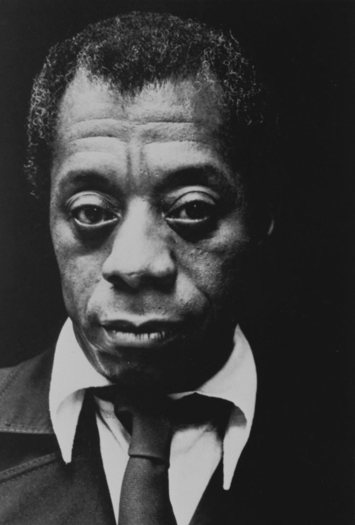 James Baldwin in a black suit with tie and a white shirt looks directly at us.