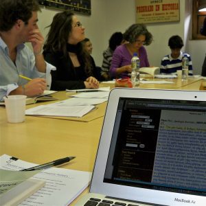 A square image with a group of people sitting around a seminar table, talking. The picture is taken from the perspective of someone sitting at the table, with the computer on the right, part of its screen visible with Greek text from the Perseus website. There are students listening to each other with papers and notebooks in front of them.