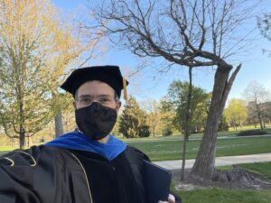 Chris Long in graduation regalia and mask in front of a leafless Resilient Tree.