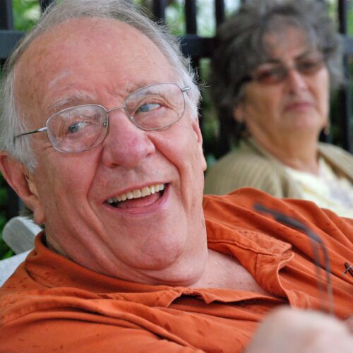 Ted Loder looking to his right with a joyful smile on his face, with Jan Filing, his wife looking on from behind and out of focus.