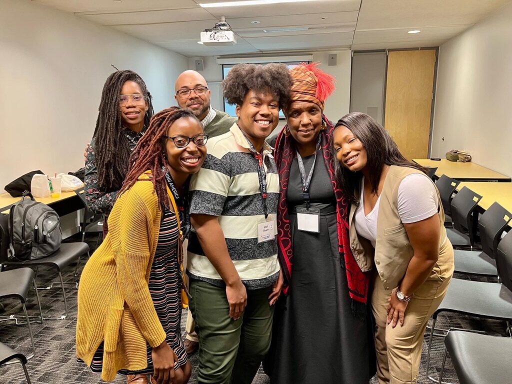 The group is gathered around Queen Cherice Harrison-Nelson who is on the right, with Toni Gordon leaning in next to her, Ural Grant on her right, Jessica Reed next to Ural, and in the back, Sharieka Botex and Marquis Taylor. They are in a conference room where the presentation on prioritizing joy in graduate education was given.