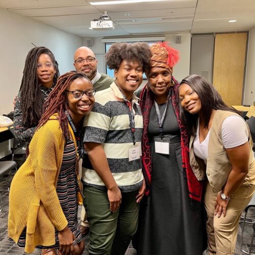 The group is gathered around Queen Cherice Harrison-Nelson who is on the right, with Toni Gordon leaning in next to her, Ural Grant on her right, Jessica Reed next to Ural, and in the back, Sharieka Botex and Marquis Taylor. They are in a conference room where the presentation on prioritizing joy in graduate education was given.