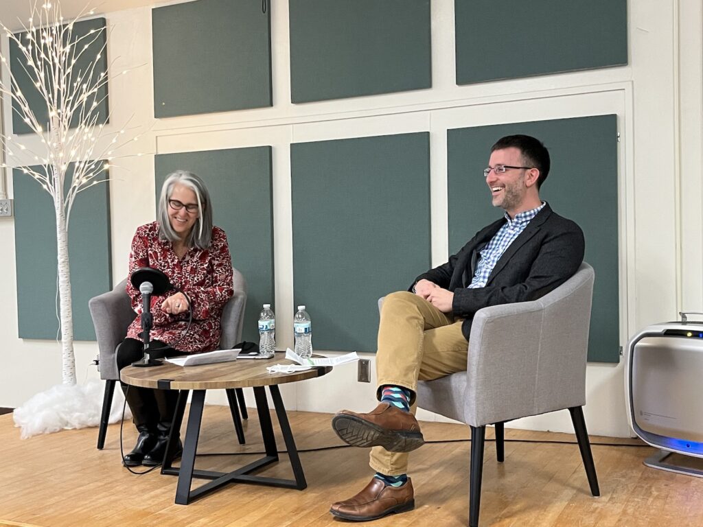 Two people sit in gray chairs with dark wooden legs on a small stage with wooden floors. There is a round coffee table made of dark and light wood between them on which is a microphone with a pop filter, papers to which the speakers are referring, and two bottles of water. Amy DeRogatis is on the left, speaking and smiling slightly as she looks down at her paper and talks into the microphone. On the right is Isaac Weiner, grinning broadly as if laughing. On the left of the stage is a fake white tree with cotton "snow" underneath and white lights on the branches. The wall behind the stage is white, broken up with gray/blue squares of fabric designed to dampen the sound. The microphone is attached by a wire that goes to an amplifier that is just visible on the right, behind Isaac. There is a blue light coming out of the bottom of the amplifier.