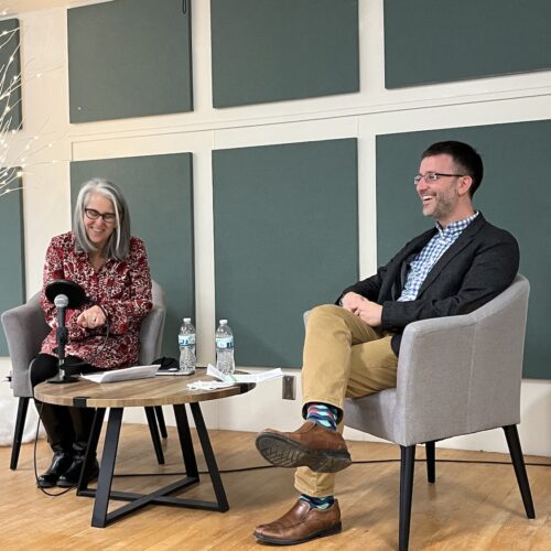 Two people sit in gray chairs with dark wooden legs on a small stage with wooden floors. There is a round coffee table made of dark and light wood between them on which is a microphone with a pop filter, papers to which the speakers are referring, and two bottles of water. Amy DeRogatis is on the left, speaking and smiling slightly as she looks down at her paper and talks into the microphone. On the right is Isaac Weiner, grinning broadly as if laughing. On the left of the stage is a fake white tree with cotton "snow" underneath and white lights on the branches. The wall behind the stage is white, broken up with gray/blue squares of fabric designed to dampen the sound. The microphone is attached by a wire that goes to an amplifier that is just visible on the right, behind Isaac. There is a blue light coming out of the bottom of the amplifier.