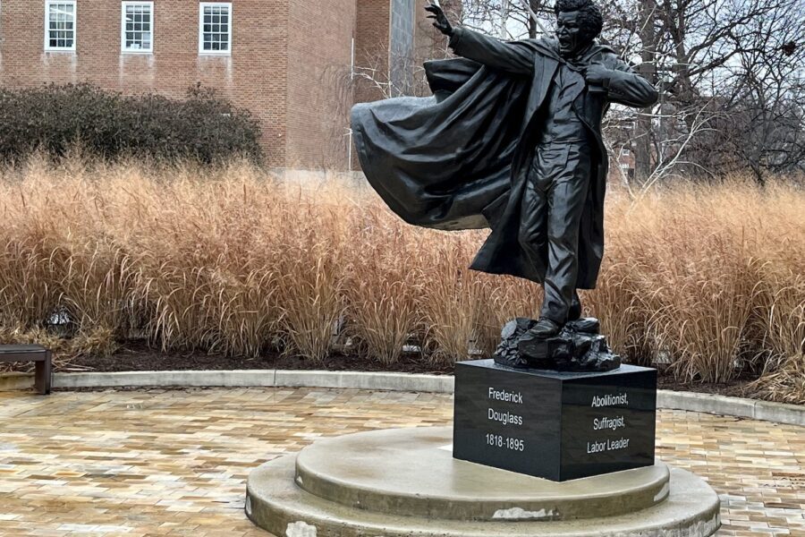 The Frederick Douglas statue on the University of Maryland campus depicts, as wikipedia puts it, "Douglass in the middle of a speech, with one arm outstretched, and a copy of his autobiography under the other arm." His mouth is open in a fiery speech. The statue is cast bronze on a black marble block with white text chiseled on front reading Frederick Douglas 1818-1895, on the side, white text is legible that reads: Abolitionist, Suffragist, Labor Leader. The figure is set off to the right of the image with a band of long, wheat colored grasses in the middle background, with three windows of the red brick colonial style building in the distant background.