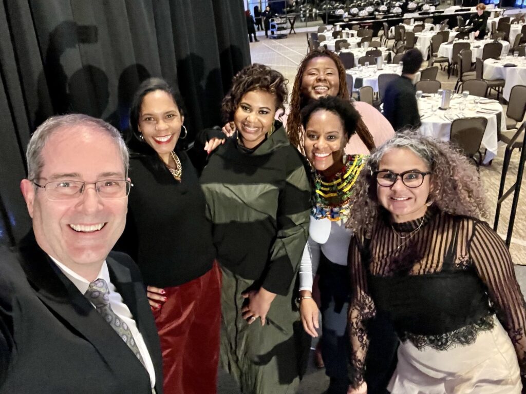 Selfie on stage at the Kellogg Center after the 43rd MLK Dinner at which LeConté Dill gave the keynote. The selfie is taken by Chris Long, who is on the left, with, from left to right, Ruth Nicole Brown, LeConté Dill, Chamara Kwakye, Gianina Strother, and Yvonne Morris. In the background are rows of empty round tables with white tablecloths and black chairs around them.