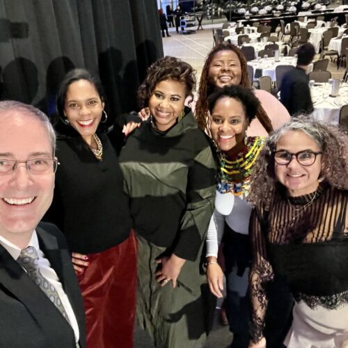 Selfie on stage at the Kellogg Center after the 43rd MLK Dinner at which LeConté Dill gave the keynote. The selfie is taken by Chris Long, who is on the left, with, from left to right, Ruth Nicole Brown, LeConté Dill, Chamara Kwakye, Gianina Strother, and Yvonne Morris. In the background are rows of empty round tables with white tablecloths and black chairs around them.