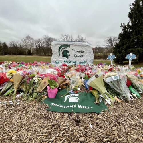 The Spartan Rock, painted white, with a green Spartan helmet, on the left and "Always a Spartan" painted on the front. The names of Brian Fraser, Arielle Anderson, and Alexandria Verner, are written in green letters along the bottom. In front of the rock are piles of flowers and a green and white Spartans Will flag laying on mulch front and center of the image. There are three crosses with hearts and fish symbols to the right of the rock.