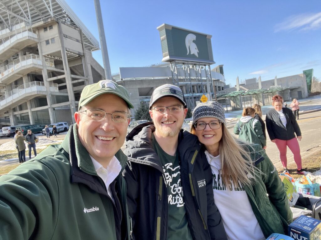 Selfie with Chris Long in a Spartan hat and jacket on the left, Aaron, and Honors College student in the middle, and Val Long in Spartan green and white on the left. In the background is Spartan Stadium with the Spartan logo large against a bright blue sky on Spartan Sunday to welcome students to campus after the February 13, 2023 shootings.