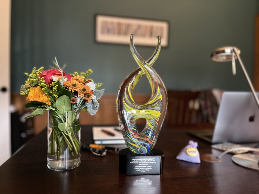 A dark wooden desktop has a bouquet of orange, white, and green flowers in a glass cylindrical vase on the left. In the middle is a multi-colored glass trophy in a double helix style on a black base on which the words are inscribed: "Michigan State University Institutional Champion Award for Community Engagement Scholarship Presented to Christopher P. Long, College of Arts and Letters and Honors College 2023." To the right, there is a small lavender bag of lavender with a yellow AAAS button on it. Blurred in the background on the right is an Apple MacBook Pro, the arm of a desk light and behind the trophy and flower are four fountain pens, a journal and a notepad.