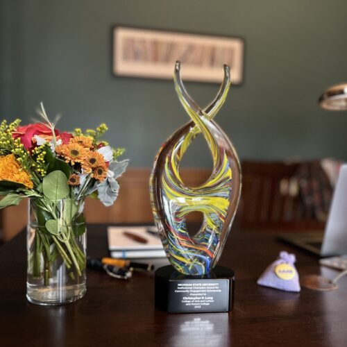 A dark wooden desktop has a bouquet of orange, white, and green flowers in a glass cylindrical vase on the left. In the middle is a multi-colored glass trophy in a double helix style on a black base on which the words are inscribed: "Michigan State University Institutional Champion Award for Community Engagement Scholarship Presented to Christopher P. Long, College of Arts and Letters and Honors College 2023." To the right, there is a small lavender bag of lavender with a yellow AAAS button on it. Blurred in the background on the right is an Apple MacBook Pro, the arm of a desk light and behind the trophy and flower are four fountain pens, a journal and a notepad.