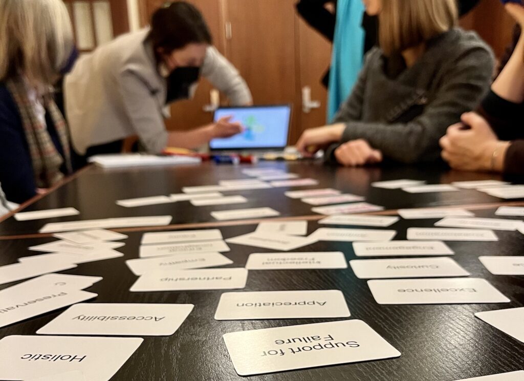 In the foreground in focus are white values cards with black letters laying on a dark wooden table with words like "Support for Failure," "Holistic," "Accessibility,"Excellence," "Appreciation," written on them. We read the cards upside-down as we look down the table, our perspective at table-level looking toward a woman in a black mask leaning over a computer pointing to something as participants around the table look toward the laptop on the table. The laptop and the people are out of focus at the depth of field is short and the focus is on the cards nearest us on the table.