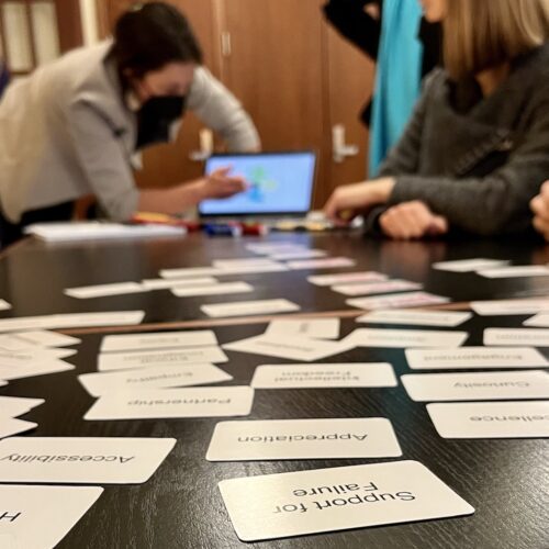 In the foreground in focus are white values cards with black letters laying on a dark wooden table with words like "Support for Failure," "Holistic," "Accessibility,"Excellence," "Appreciation," written on them. We read the cards upside-down as we look down the table, our perspective at table-level looking toward a woman in a black mask leaning over a computer pointing to something as participants around the table look toward the laptop on the table. The laptop and the people are out of focus at the depth of field is short and the focus is on the cards nearest us on the table.