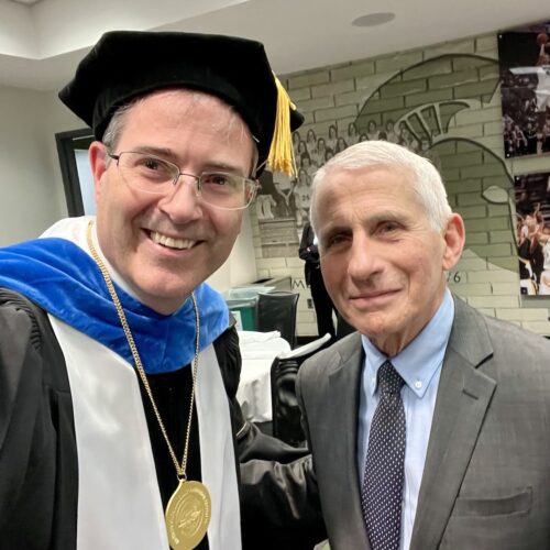 A selfie with Chris Long in academic regalia to the right of Dr. Anthony Fauci in the Breslin Center before the 2023 MSU Doctoral graduation ceremony. The wall behind them has a Spartan helmet and images of Spartan Basketball players in action.