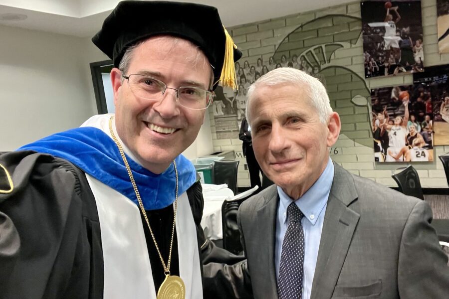 A selfie with Chris Long in academic regalia to the right of Dr. Anthony Fauci in the Breslin Center before the 2023 MSU Doctoral graduation ceremony. The wall behind them has a Spartan helmet and images of Spartan Basketball players in action.