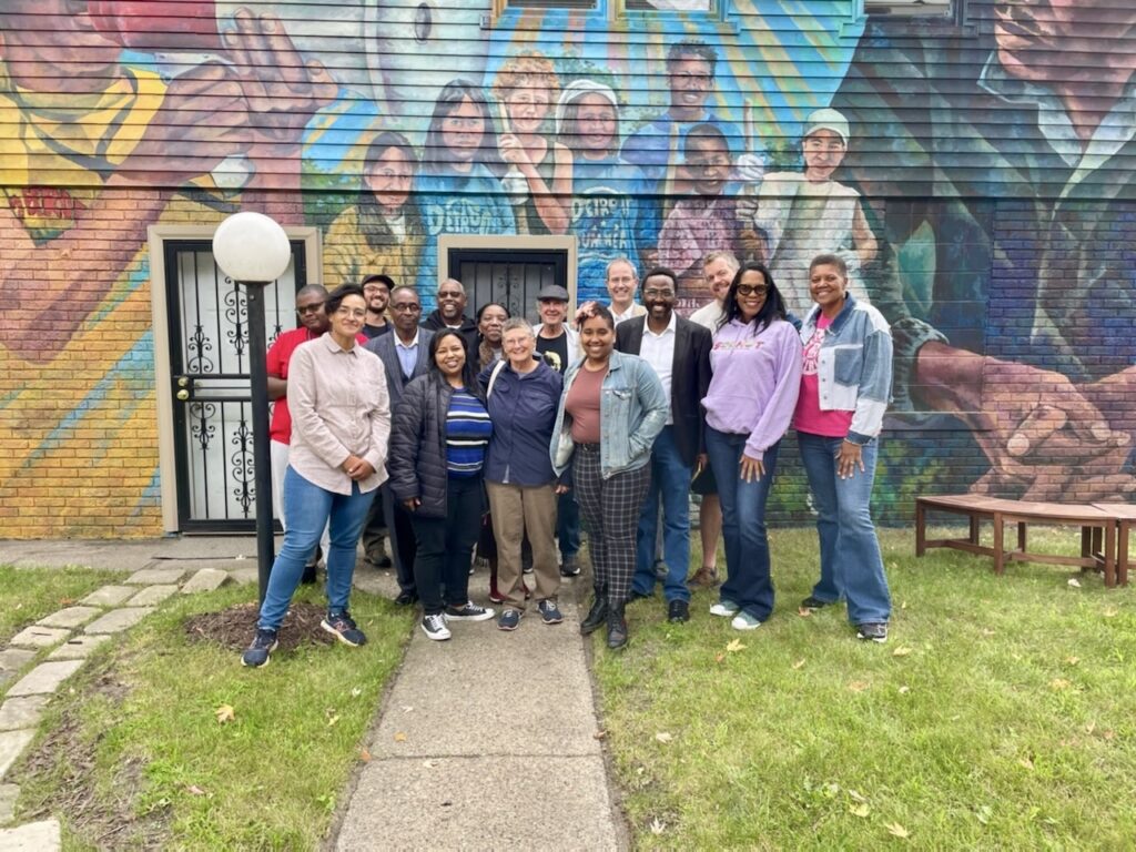 A diverse group of people of wide-ranging ages stand in a courtyard in front of a colorful mural of Grace Lee and James Boggs with young people. A young Grace Lee Boggs is in yellow, holding a megaphone to her mouth. Jimmie Boggs, on the right, is in a dark jacket, hands folded in front of me, listening. There is a diverse group of young people between them.