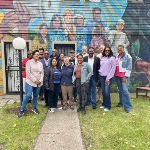 A diverse group of people of wide-ranging ages stand in a courtyard in front of a colorful mural of Grace Lee and James Boggs with young people. A young Grace Lee Boggs is in yellow, holding a megaphone to her mouth. Jimmie Boggs, on the right, is in a dark jacket, hands folded in front of me, listening. There is a diverse group of young people between them.