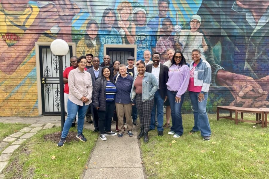 A diverse group of people of wide-ranging ages stand in a courtyard in front of a colorful mural of Grace Lee and James Boggs with young people. A young Grace Lee Boggs is in yellow, holding a megaphone to her mouth. Jimmie Boggs, on the right, is in a dark jacket, hands folded in front of me, listening. There is a diverse group of young people between them.