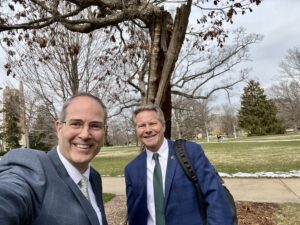 Selfie with Chris Long and Kevin Guskiewicz in front of the Resilient Tree on MSU's campus. Long and Guskiewicz are in suit jackets with green ties and the Beaumont Tower is off in the distance on the left. The gnarled branches of the tree stretch out above them.