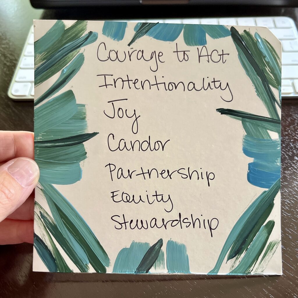 A square card painted by Katy Krieger with teal and green paint strokes along the edges with the values of the Provost's Office listed: Courage to Act, Intentionality, Joy, Candor, Partnership, Equity, Stewardship.