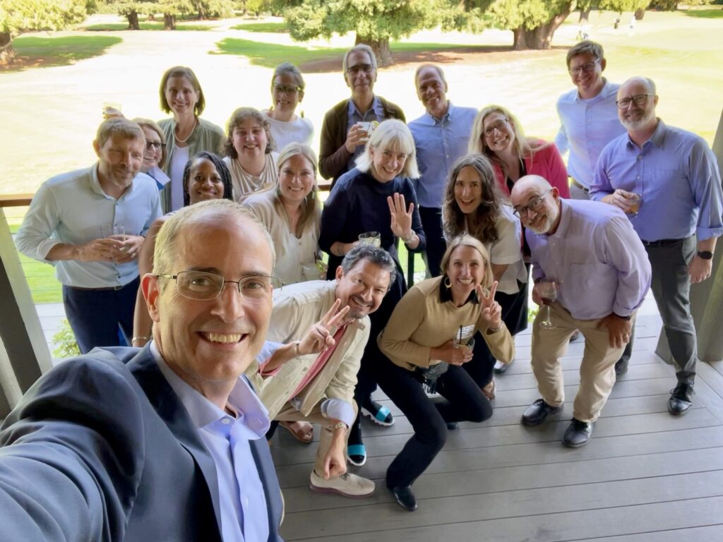 Selfie with Provost Long and members of the Provost's Council smiling and waving and giving the peace sign on a porch in front of a lawn with trees in the background.