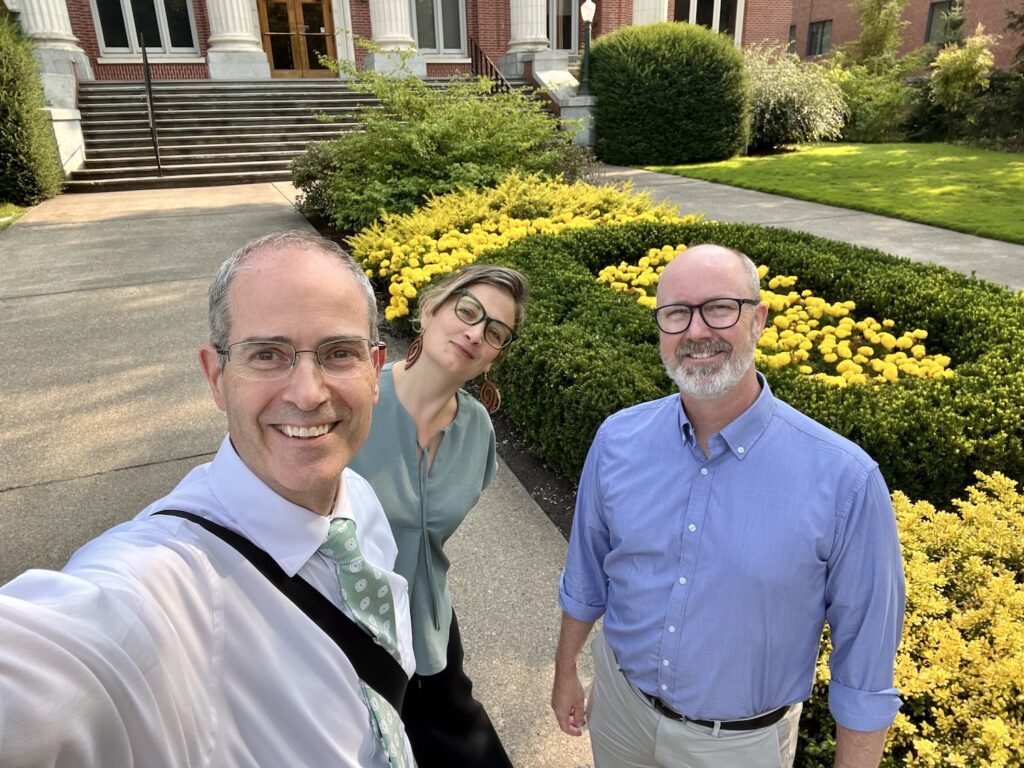 A selfie with Chris Long, Nicky Agate, and Jason Rhody in front of Johnson Hall at the University of Oregon with a hedge in the shape of an "O" and yellow flowers in the middle.