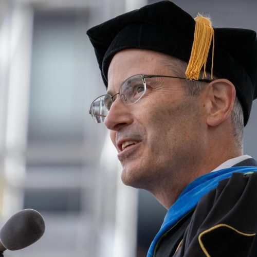 Provost Long in academic regalia, including a black cap with a yellow tassel, speaks at a podium during a graduation ceremony.