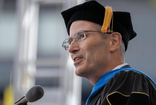 Provost Long in academic regalia, including a black cap with a yellow tassel, speaks at a podium during a graduation ceremony.