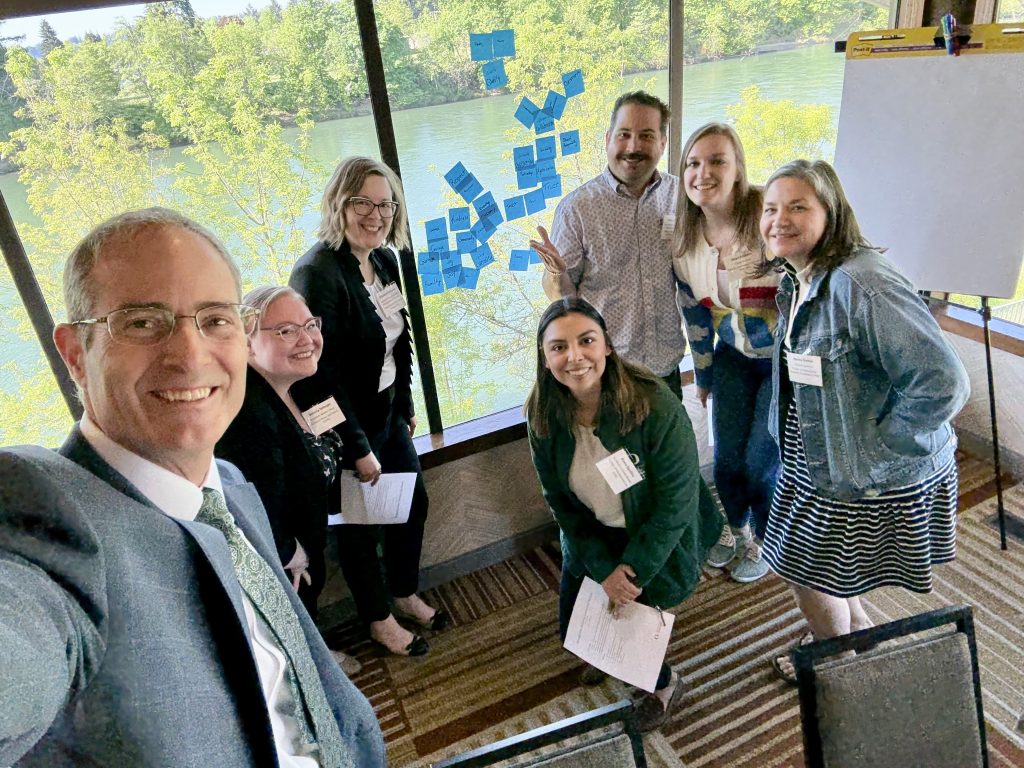 Provost Long with six University of Oregon leaders in advising in business attire smile for a group selfie in a meeting room with large windows, trees and water outside. Sticky notes cover part of the window, and a blank flip chart stands nearby.