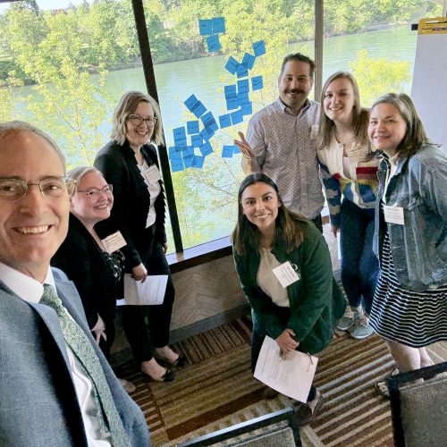 Provost Long with six University of Oregon leaders in advising in business attire smile for a group selfie in a meeting room with large windows, trees and water outside. Sticky notes cover part of the window, and a blank flip chart stands nearby.