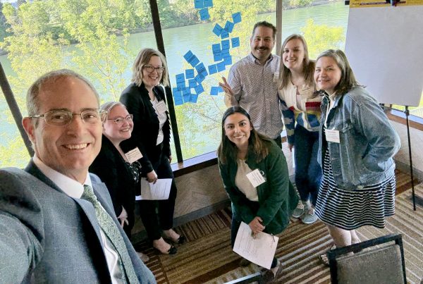 Provost Long with six University of Oregon leaders in advising in business attire smile for a group selfie in a meeting room with large windows, trees and water outside. Sticky notes cover part of the window, and a blank flip chart stands nearby.