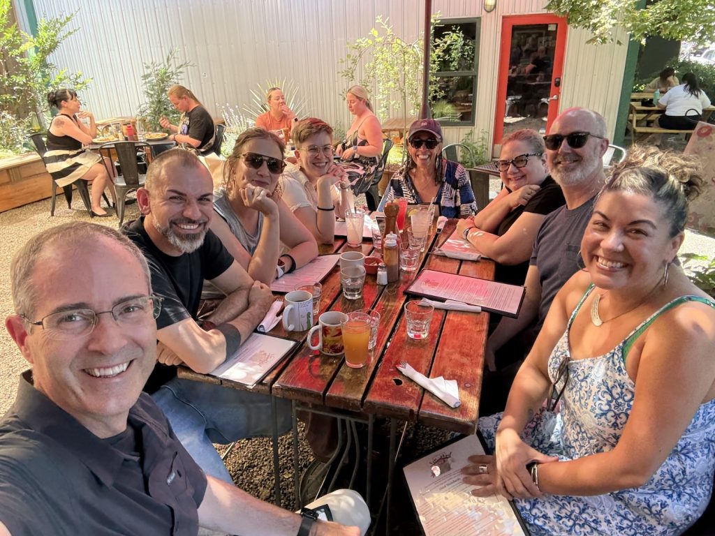 A group of nine adults sit around a red outdoor table at a café, smiling at the camera. The table has drinks, mugs, and condiments, and the setting is sunny with greenery and other diners in the background.