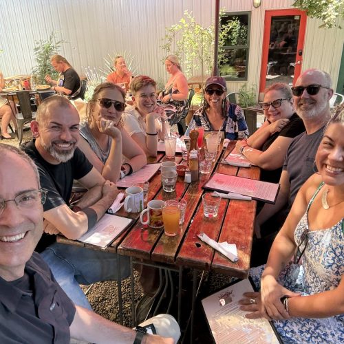 A group of nine adults sit around a red outdoor table at a café, smiling at the camera. The table has drinks, mugs, and condiments, and the setting is sunny with greenery and other diners in the background.