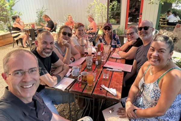A group of nine adults sit around a red outdoor table at a café, smiling at the camera. The table has drinks, mugs, and condiments, and the setting is sunny with greenery and other diners in the background.