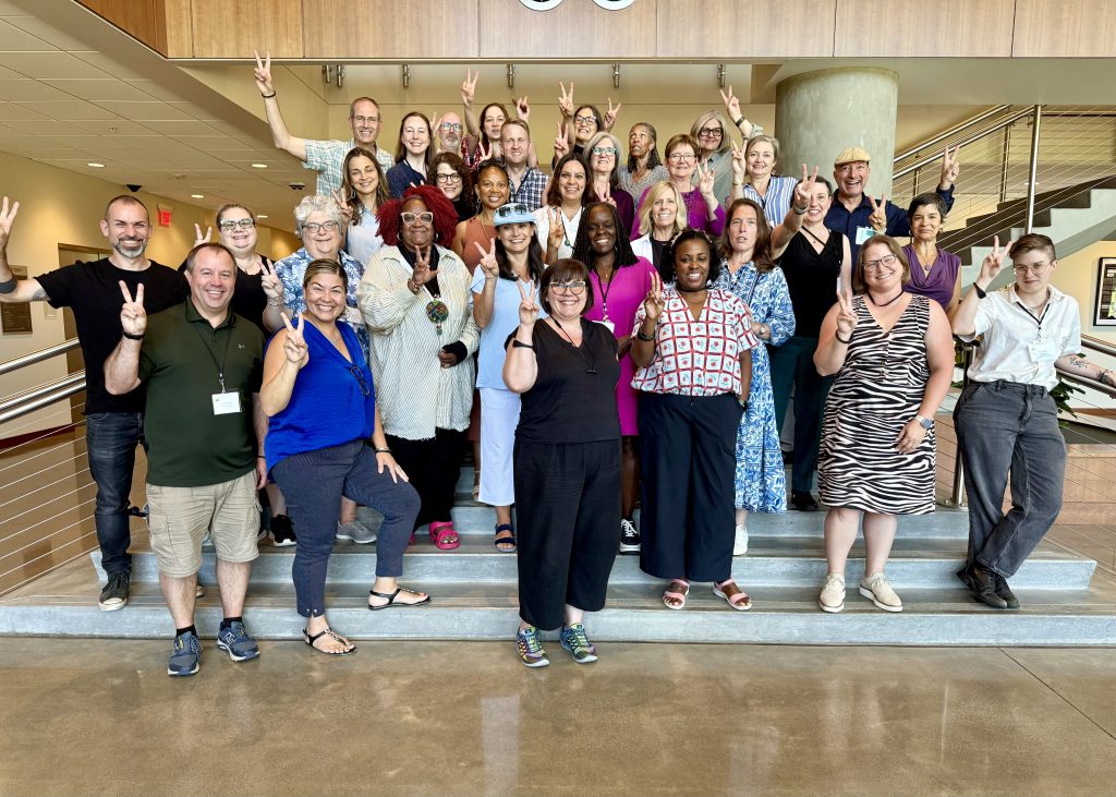 A diverse group of people pose together on indoor steps, smiling and holding up peace signs, or "V" signs for VELI. Some stand, while others kneel or sit. The background shows a modern interior with wood accents and metal railings.