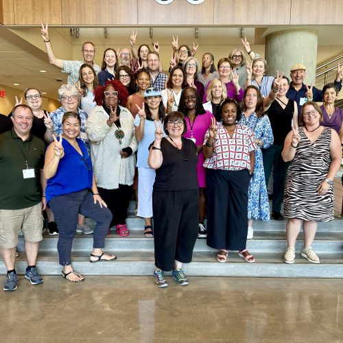 A diverse group of people pose together on indoor steps, smiling and holding up peace signs, or "V" signs for VELI. Some stand, while others kneel or sit. The background shows a modern interior with wood accents and metal railings.