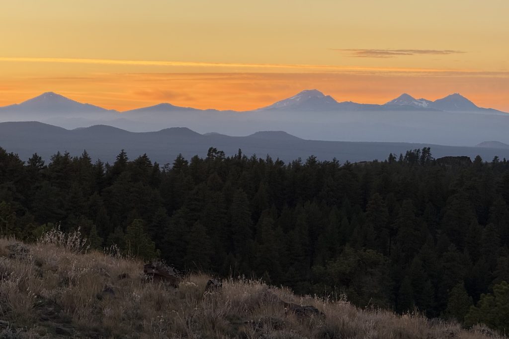 Sunset over the distant snow-capped mountains of Sisters and Mt. Batchelor in Oregon with orange and yellow sky, dark forest in the middle ground, and the dry grassy hillside of Pine Mountain in the foreground.