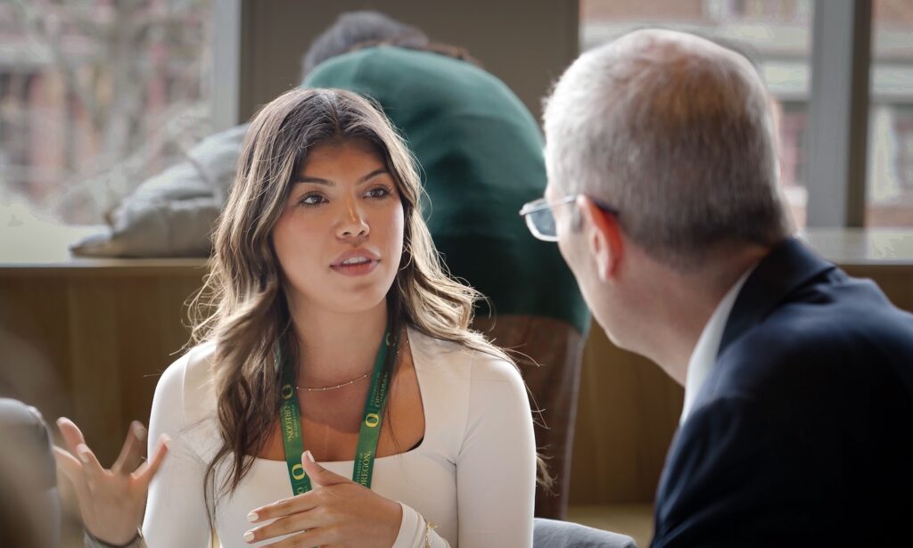 A woman with long brown hair, wearing a white top and a green lanyard, gestures while speaking to a man in glasses and a suit. They are seated indoors, with large windows and blurred figures in the background.