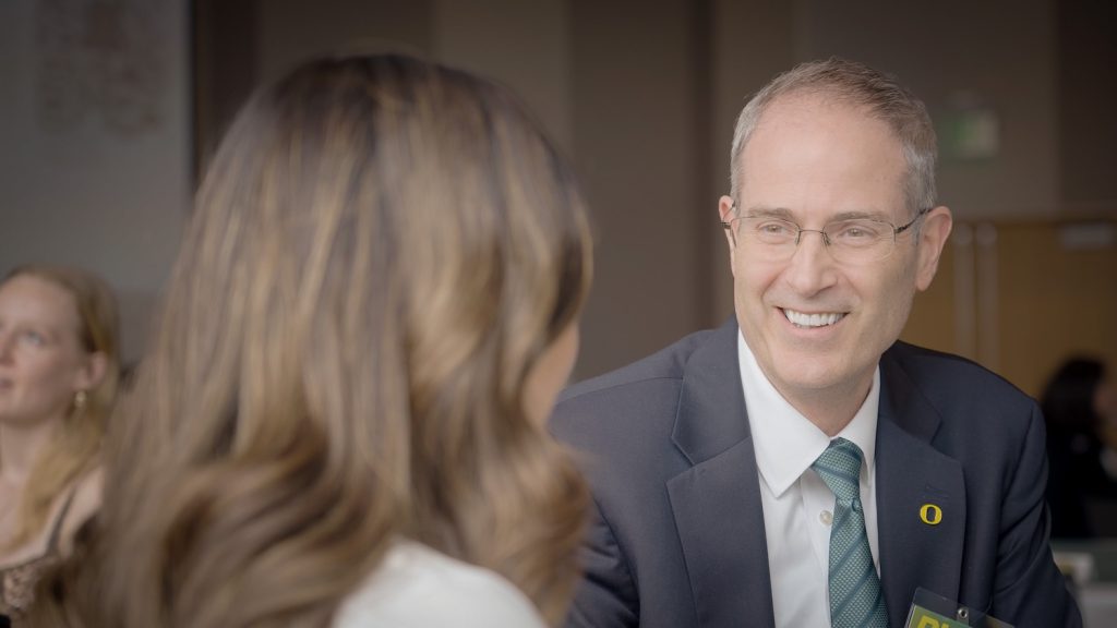 Provost Long in a suit and green tie with an O pin is smiling and talking to a prospective student with long brown hair. They are indoors at what appears to be a formal or professional event. Other people are blurred in the background.