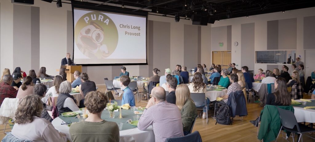 A large group of people sits at round tables in a spacious room, listening to a speaker at a podium. A screen displays PURA and Chris Long Provost along with a graphic of a Oregon Duck with a magnifying glass.