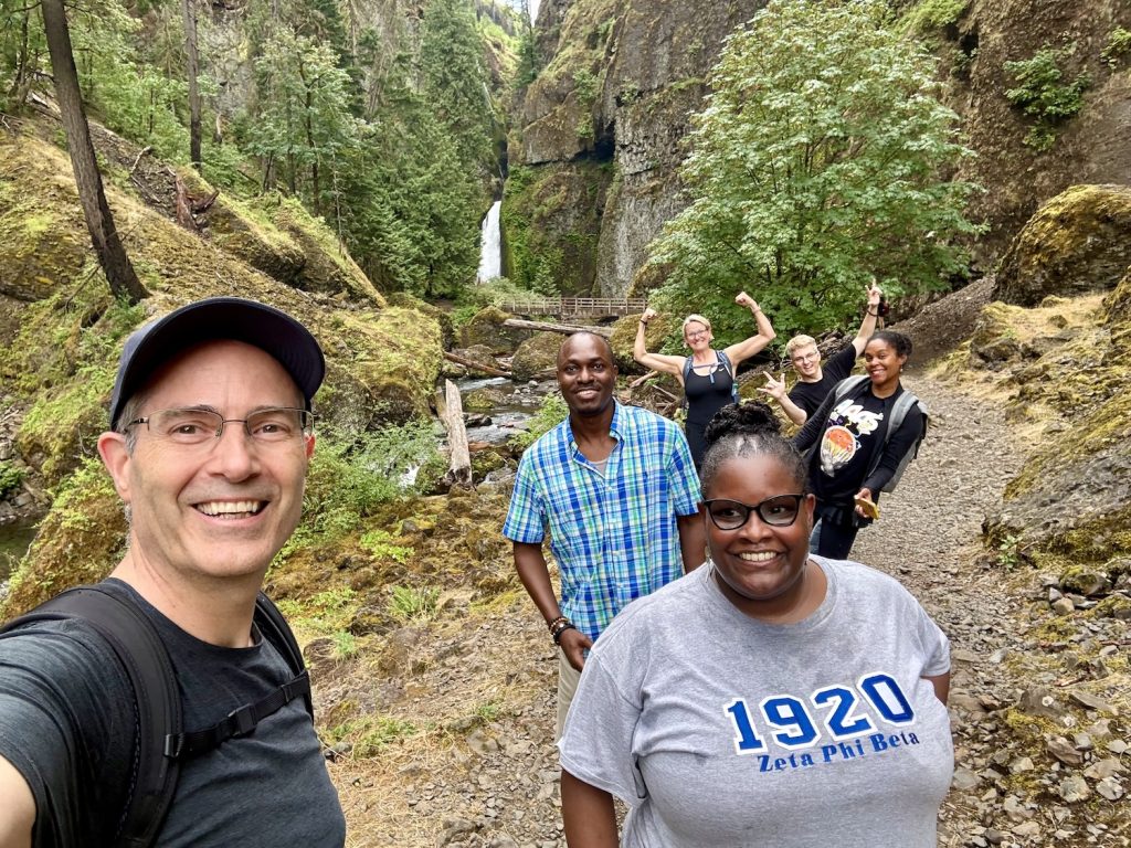 A group selfie on a forested trail with a tall waterfall in the background, cascading down a mossy rock face. A man in a cap and glasses takes the photo in the foreground; six other hikers stand behind him, several flashing peace signs, one woman wearing a gray "1920 Zeta Phi Beta" t-shirt.