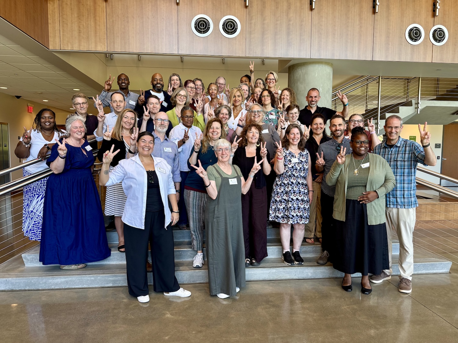 A large group of about 30 people posed on an indoor staircase with wood-paneled walls, most making "V" for VELI signs and smiling for a group photo.