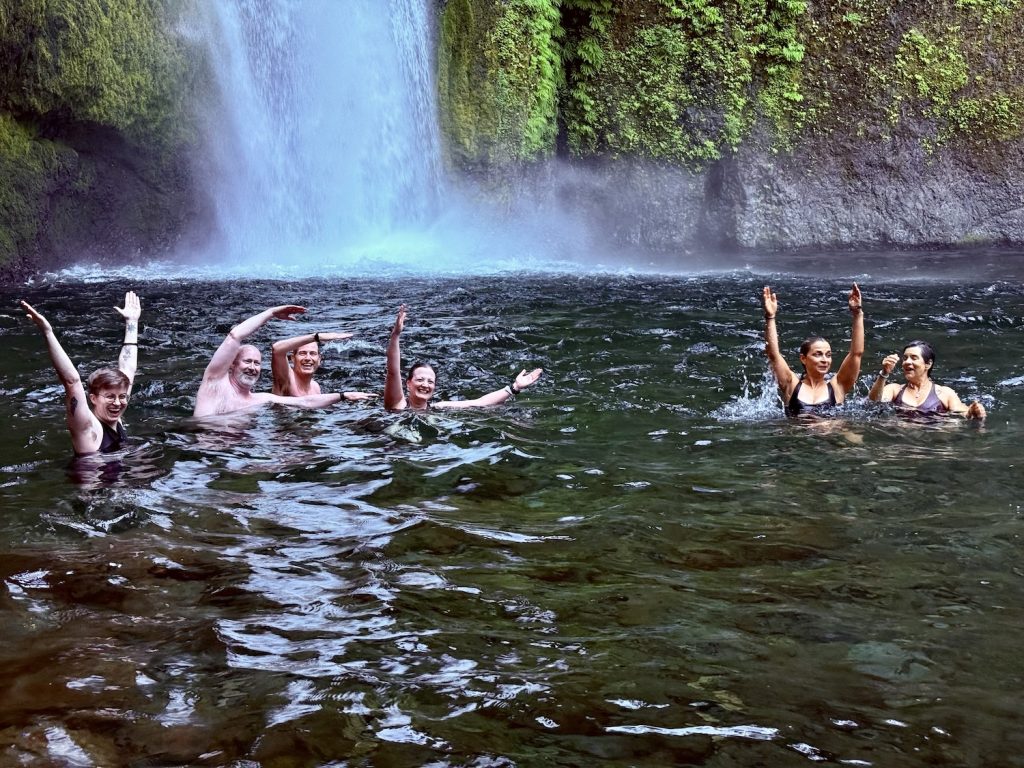 Six people swimming and trying to spell out "VELI" in a pool at the base of Wahclella Falls, with a wide curtain of water falling over a moss-covered rock wall behind them.