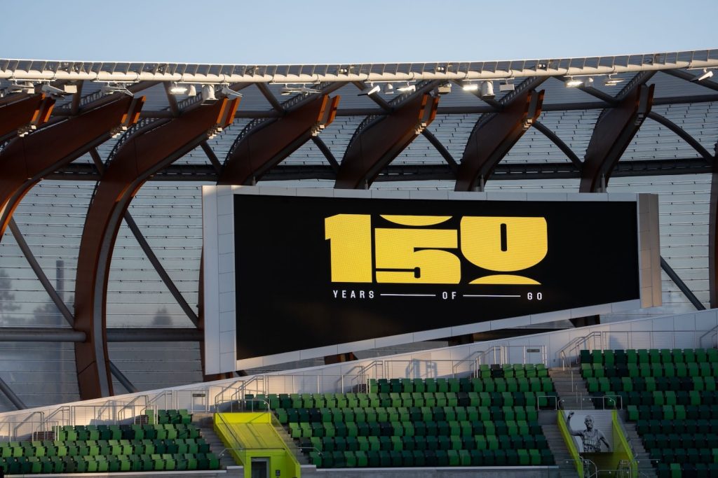 A large video board hangs beneath the curved timber-and-steel roof of a Hayward Field at golden hour. It shows a bold gold "150" with the words "YEARS — OF — GO" beneath it on a black background. Rows of green seats fill the lower part of the frame. A black-and-white portrait of an athlete is partly visible near the lower right.