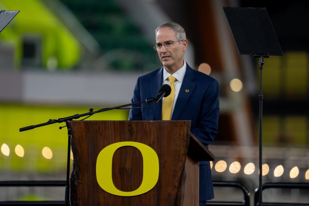 Provost Long in glasses, a navy suit and a gold tie, with a small yellow "O" pin on his lapel, speaks into a microphone at a live-edge wooden podium. The podium carries a large yellow University of Oregon "O." A teleprompter stands to his right. Behind him, the stadium is blurred, with bright green seating and strings of warm lights.